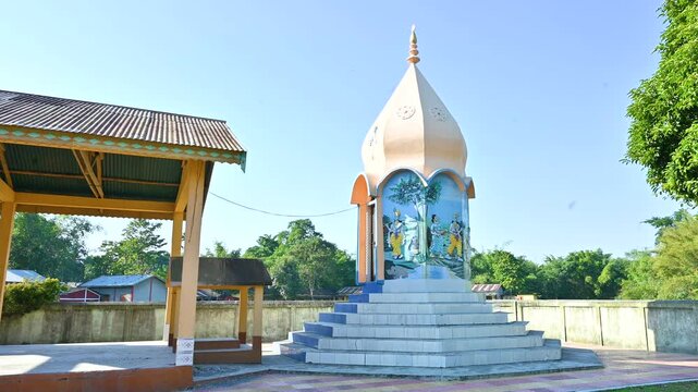 A compact temple with dome, artwork, and nearby pavilion stands among trees in Byaskuchi village, Assam. Captures rural spirituality and architecture.