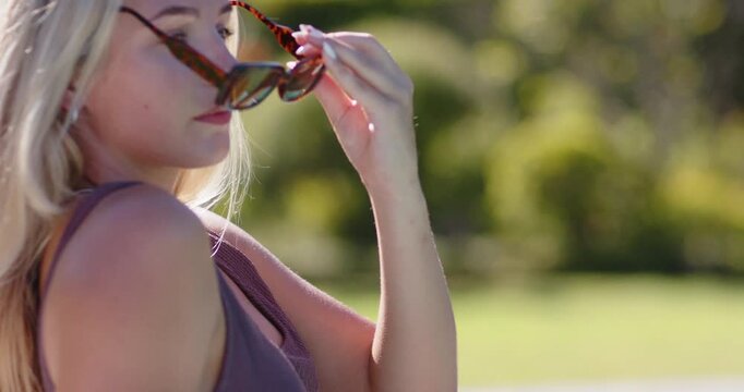 Vertical video: Woman 20s lifting tortoise sunglasses poolside, sun causing squinting and adjusting