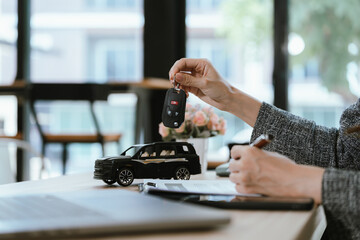 Fototapeta premium A hand holding a car key above a toy car on a desk, symbolizing car purchase, insurance, ownership, or dealership transaction in a modern office setting.