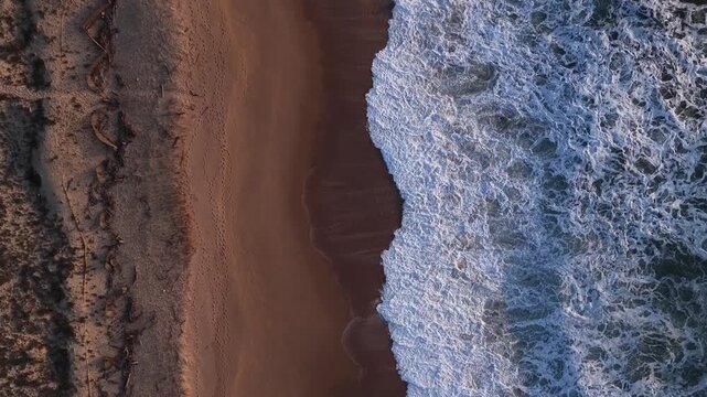 Aerial view of the textured coastline where foamy waves crash onto the sandy beach, creating a mesmerizing contrast of colors, Lisbon, Portugal.