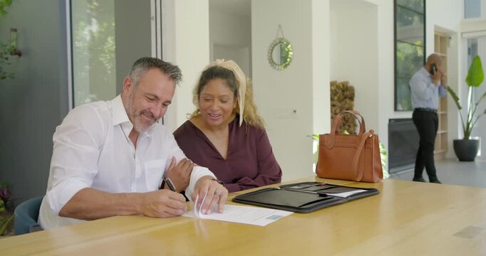 After agent call, Diverse couple sitting at table in modern home signing documents finalizing deal
