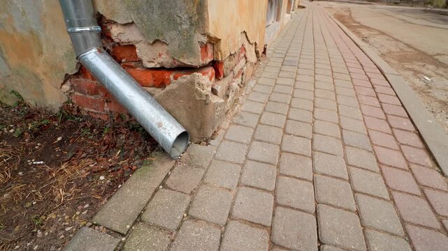Broken downspout drains onto a sidewalk beside exposed brick and crumbling plaster, showing poor drainage and a weathered facade on an old urban building.