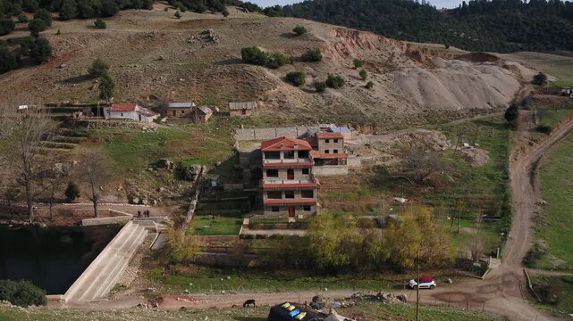 A cinematic 4K aerial wide shot of a serene mountain lake and a small dam in the Moroccan Middle Atlas, featuring traditional guest houses and parked cars on the shore, surrounded by dense evergreen f