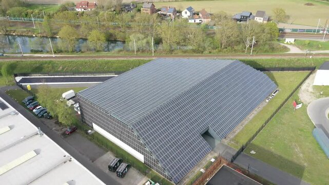 Aerial view of datacenter covered in solar panels, juxtaposed against green grass, a canal, and railway lines, Oostkamp, Flanders, Belgium.