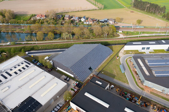 Aerial view of a datacenter covered with solar panels juxtaposed with a canal, railway tracks, and small houses in the background, Oostkamp, Flanders, Belgium.