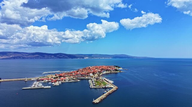 Nesebar Old Town shows buildings near the water with boats in the harbor. The scene has blue water, clouds in the sky, and a sunny day. People walk along the coast.