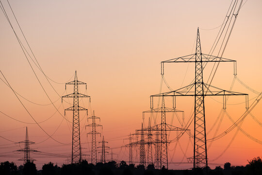 Silhouette of several high-voltage power lines and electricity pylons against a glowing orange and red sunset sky in Germany.
