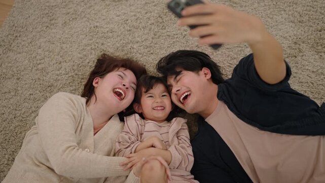 Happy Asian family lying on carpet and taking selfie with smartphone