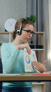 Close up of good-looking satisfied skilled young girl in headset which sitting at her workplace at home and presenting her statistics datas with charts to mentor or tutor via video meeting