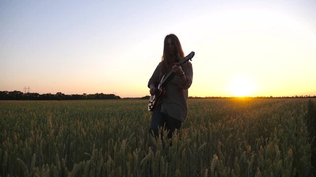 Hippie man with long hair plays on electric guitar walking through green barley meadow. Male hipster going among wheat field and playing on musical instrument. Beautiful sunset at background. Slow mo