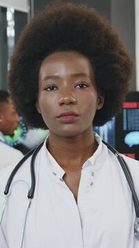 Pretty concentrated purposeful coalified female african american doctor in white gown looking at camera with strict face on the background of multiethnic colleagues which talking near digital