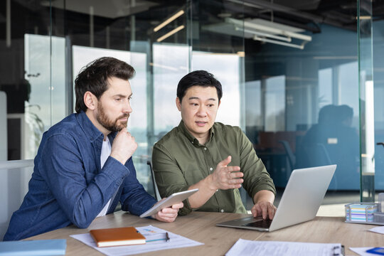 Two diverse business professionals collaborating during a discussion, reviewing information on a laptop and tablet, working together on a project in a bright contemporary office