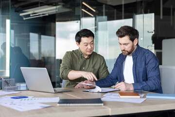 Obraz premium Two diverse businessmen are analyzing documents and discussing details around a desk in a modern office, working together on a strategic partnership or agreement