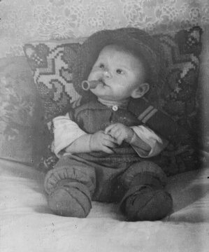 Infant with pacifier in mouth sitting propped against embroidered decorative pillow in knit outfit, scanned black and white print circa 1950s