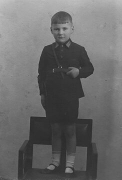 Young boy in dark school jacket with shoulder bag standing on wooden chair for studio portrait, scanned black and white print circa 1940s