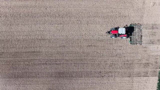 Aerial view of a red tractor pulling a harrow across a vast field of tilled soil, creating a dusty trail, Wintzenheim-Kochersberg, Grand Est, France.