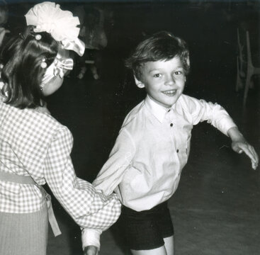 Boy and girl dancing together at indoor school event, boy smiling with arms outstretched, girl in checkered dress with bow, scanned black and white print circa 1960s