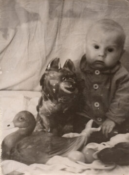 Young toddler in button jacket sitting with decorative ceramic horse and duck toy figurine, blurry sepia archival print circa 1930s