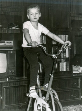 Young boy in white t-shirt sitting on stationary exercise bicycle in living room with bookshelf and vintage television, scanned black and white print circa 1980s