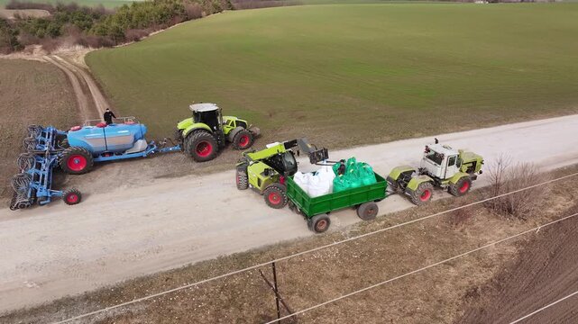 Ternopil, Ukraine - March 25, 2026: Agricultural machinery in operation, featuring a green telehandler lifting a blue tank and a large plow on a farm field with brown soil and green grass