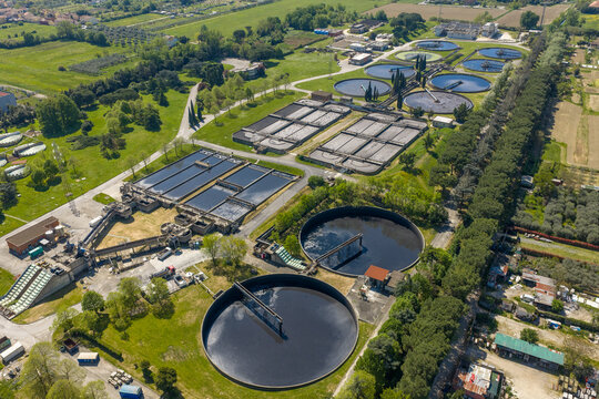Aerial view of a large water treatment plant with circular sedimentation tanks and rectangular basins surrounded by green fields and trees in Prato, Tuscany, Italy.