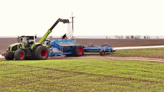 Ternopil, Ukraine - March 25, 2026: Green telehandler lifts a blue tank while a large plow is attached, set in a farm field with brown soil and green grass in the background