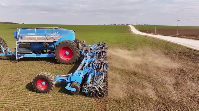 Green telehandler lifts a blue tank while a large plow is attached, set in a farm field with brown soil and green grass in the background