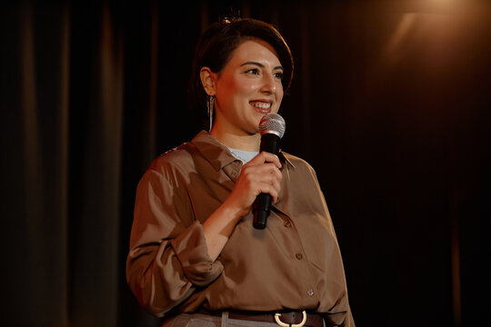 Medium shot of biracial young adult woman performing standup comedy, speaking into microphone on stage. Suitable for entertainment marketing, live event promotion, public speaking themes
