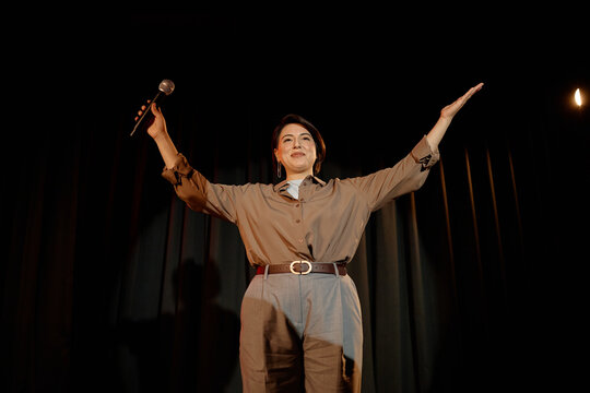 Biracial young adult woman performing standup on stage, raising arms and holding microphone to engaging audience. Ideal for comedy promotion, live event marketing, entertainment branding
