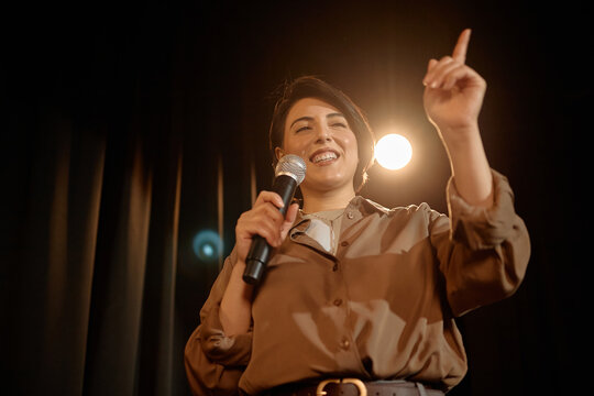 Young adult woman performing standup on stage, speaking into microphone and gesturing toward audience. Ideal for comedy promotion, live event marketing, entertainment advertising
