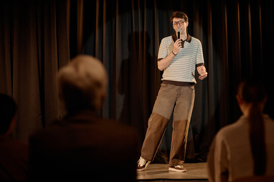 Young adult Caucasian man performing standup comedy on stage, speaking into microphone before audience. Useful for entertainment marketing, live event promotion, medium full shot
