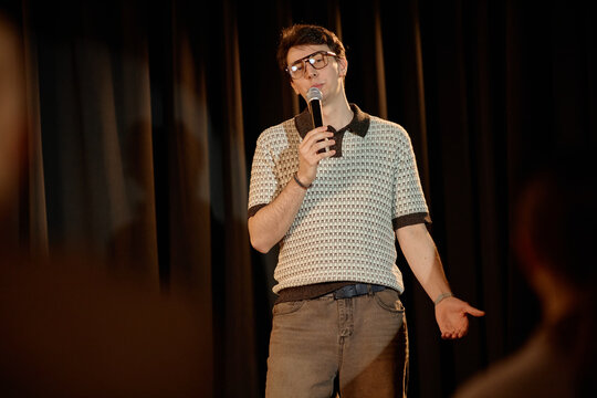Young adult Caucasian man performing standup comedy on stage, speaking into microphone and engaging audience. Useful for entertainment marketing, live event promotion, medium shot
