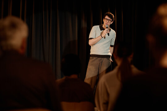 Young adult Caucasian man performing standup comedy on stage, speaking into microphone before audience. Useful for entertainment marketing, live event promotion, comedy club ads
