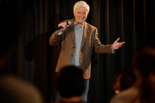 Senior Caucasian man performing standup comedy on stage, speaking into microphone and gesturing to audience. Useful for entertainment marketing, live event promotion, medium shot
