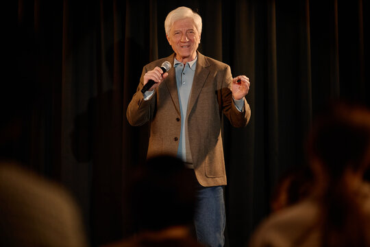 Portrait of Caucasian senior man performing standup on stage, speaking into microphone before audience, conveying live entertainment and public speaking. Ideal for comedy promotion and event marketing