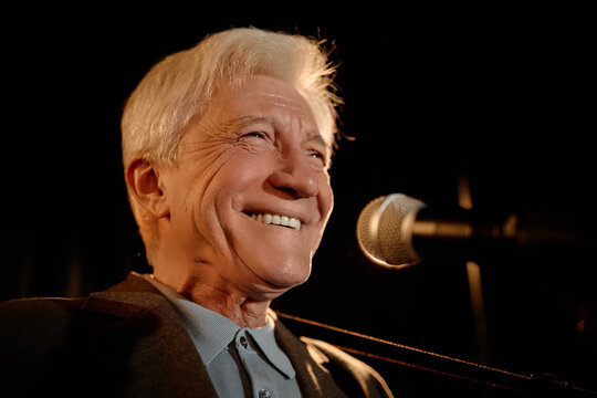 Portrait of Caucasian senior man performing standup on stage, smiling beside microphone and engaging audience. Useful for comedy promotion, live event marketing, entertainment branding
