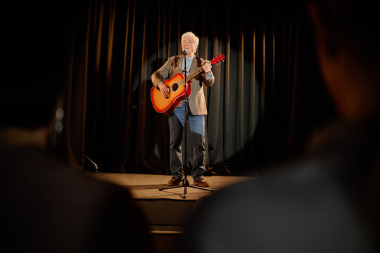 Wide shot of Caucasian senior man performing standup music set, singing into microphone and playing acoustic guitar on stage. Useful for live entertainment, talent show, event promotion
