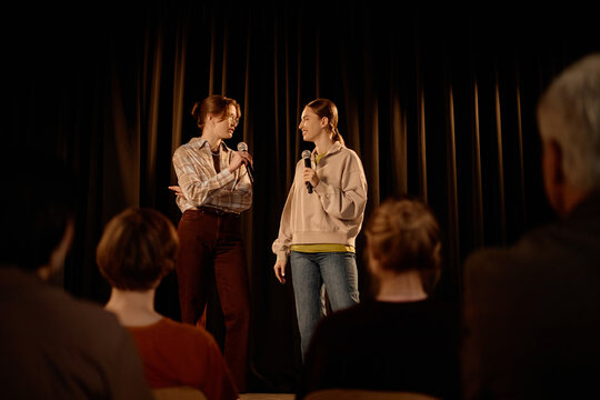 Wide shot of young adult Caucasian women performing standup dialogue on stage, speaking into microphones before audience. Useful for comedy events, live entertainment promotion, public speaking
