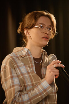 Young adult Caucasian woman performing standup on stage, speaking into microphone during live set. Useful for comedy promotion, event marketing, entertainment advertising, close up

