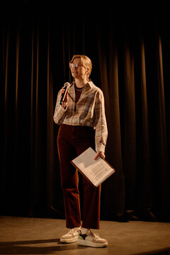 Vertical shot of young adult Caucasian woman speaking on stage with microphone and notes, presenting standup set or live event. Useful for comedy promotion, hosting, public speaking marketing
