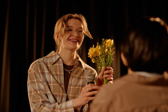 Teenage Caucasian woman receiving flowers and microphone on stage during standup performance, expressing audience appreciation and live entertainment success. Medium shot for event promotion
