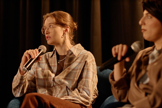 Young adult Caucasian women speaking into microphones during stage discussion panel, sharing opinions for live audience. Ideal for event marketing, public speaking, entertainment promotion
