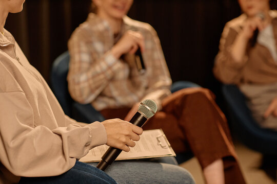 Young adult woman holding microphone and moderating standup discussion on stage with speakers in background. Useful for event promotion, panel talk, public speaking, medium shot
