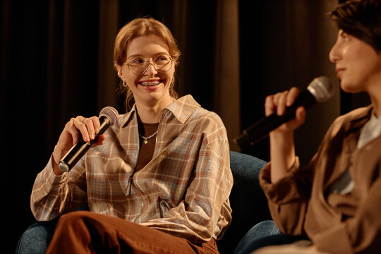 Young adult Caucasian women speaking into microphones during stage discussion, sharing ideas in live standup format. Useful for event promotion, public speaking, entertainment marketing
