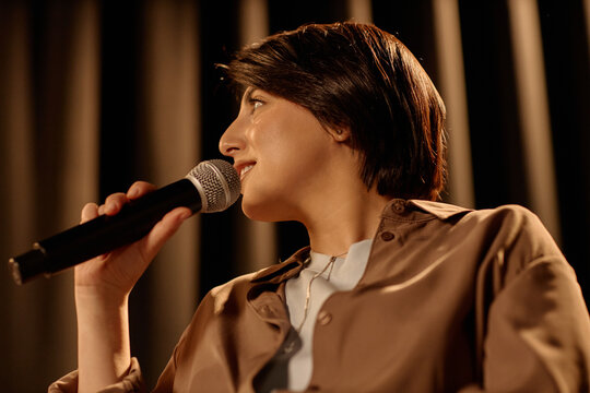 Young adult woman performing standup comedy on stage, speaking into microphone during live set. Useful for entertainment marketing, event promotion, public speaking campaigns
