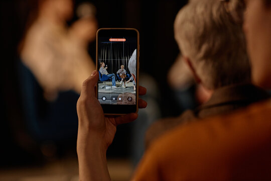 Close up of middle aged attendee recording standup talk on smartphone, capturing speakers on stage. Useful for event marketing, live entertainment promotion, social media content
