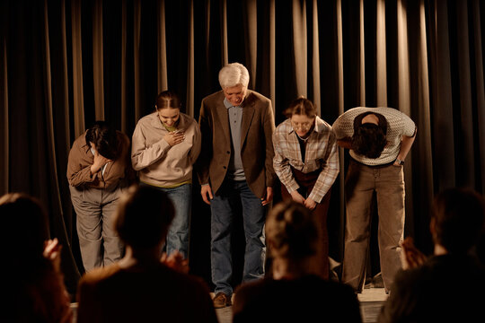 Wide shot of diverse theater cast bowing on stage after standup performance, acknowledging audience applause. Suitable for live entertainment promotion, event marketing, ticket sales
