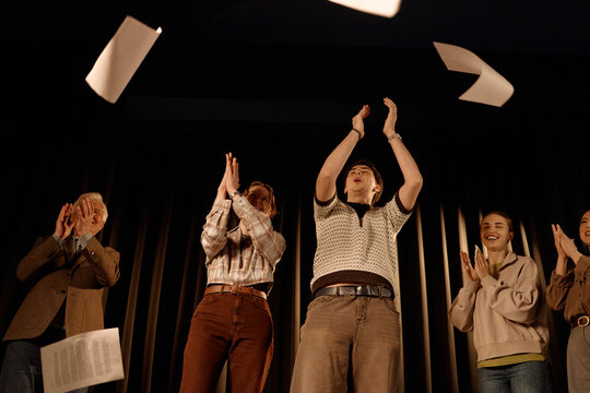 Diverse adults standing on stage, clapping and tossing papers upward during standup performance or live event. Supporting entertainment promotion, team celebration, public speaking, wide angle
