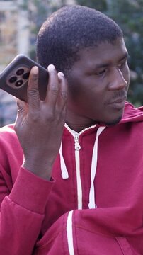 Vertical: Young African American man in a red hoodie listens to a voice message on his smartphone. His focused expression reflects attention and engagement in modern digital communication.