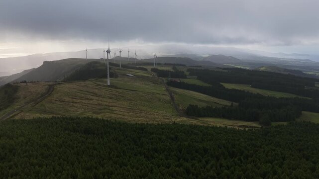 Aerial view of wind turbines standing tall on a grassy hill, contrasting with the dark green forests and cloudy skies, Ponta Delgada, Sao Miguel, Portugal.